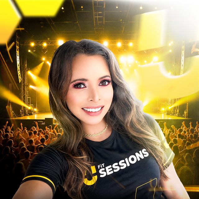 Amanda Mendes sorrindo, de camiseta preta do UFit Sessions, com o palco iluminado ao fundo